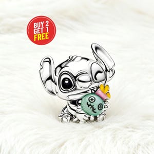May include: A silver charm depicting the character Stitch from Lilo & Stitch, winking and holding a green object with a yellow and pink accent. The charm is set against a white, fluffy background. A red circle with the text "BUY 2 GET 1 FREE" is in the upper left.
