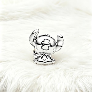 May include: A silver charm depicting the character Stitch from Disney's Lilo & Stitch. The charm is detailed with the Disney and Pandora logos. The charm is on a white, fluffy surface.