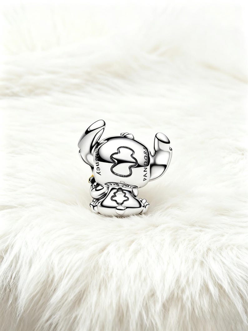 May include: A silver charm depicting the character Stitch from Disney's Lilo & Stitch. The charm is detailed with the Disney and Pandora logos. The charm is on a white, fluffy surface.
