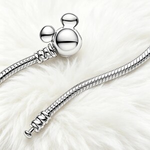 Peut inclure: Un bracelet argent&eacute; avec un fermoir en forme de Mickey Mouse. Le bracelet pr&eacute;sente un motif de cha&icirc;ne serpent. Le fermoir est compos&eacute; d'une t&ecirc;te ronde et de deux petites oreilles rondes. Le bracelet est pr&eacute;sent&eacute; sur une surface blanche et duveteuse.