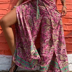Puede incluir: Pantalones envolventes de pierna ancha con un vibrante estampado en forma de abanico en tonos rosa, morado y verde oliva. La prenda incluye un cierre de lazo en la cintura y un borde ancho y contrastante en turquesa y rosa en el dobladillo.