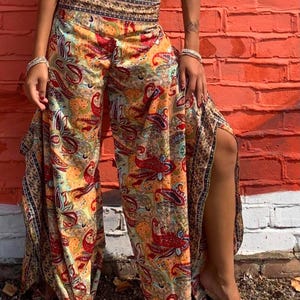 May include: Wide-leg pants featuring a vibrant, multi-colored paisley print in shades of orange, red, yellow, and blue. The pants have a high, fitted waistband with a decorative border pattern and high side slits that extend from the hem upward.
