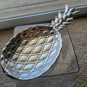 May include: Silver-colored pineapple-shaped serving dish with a diamond pattern. The dish sits on a clear plastic container, suggesting a decorative or functional item for serving or display. The pineapple design adds a tropical touch.