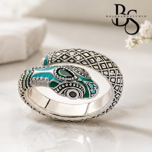 May include: A silver wrap ring shaped like a snake with a textured scale pattern. The snake head features vibrant teal and green enamel detailing, a dark teardrop-shaped stone, and a small black circular eye. The BoldBandStudio logo is visible.