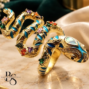 Puede incluir: Un anillo de serpiente en tono dorado con un diseño en espiral y detalles en esmalte azul brillante. El cuerpo está decorado con pequeñas piedras preciosas multicolores, incluyendo piedras rosas, verdes, naranjas y moradas.