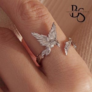 May include: A silver-toned open ring featuring a detailed bird with spread wings on one end and a leaf-like design on the other. The metal surface has a textured finish. The logo BOLD BAND STUDIO is visible in the upper right corner of the image.
