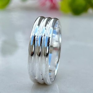 Puede incluir: Un anillo de plata con tres bandas texturizadas. El anillo está muy pulido y refleja la luz, sobre un fondo borroso de follaje verde y flores rosas. El anillo es una pieza de joyería.