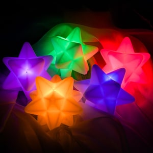 May include: A collection of five star-shaped LED lights glowing in vibrant colors including purple, green, orange, blue, and pink. Each star features a geometric, multi-faceted surface that diffuses the internal light, creating a soft, radiant effect.