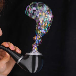 May include: A black smoking pipe with a silver-colored band emitting a large, swirling cluster of iridescent, multi-colored soap bubbles that rise upward in a curved formation against a solid black background.