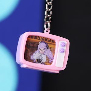以下が含まれることがあります： 銀色のチェーンが付いた、ピンクと紫色のミニチュアテレビのキーホルダー。画面には紫色の髪の毛のアニメキャラクターが表示されています。テレビの側面には紫色のボタンが2つあります。背景は青と黒のグラデーションです。
