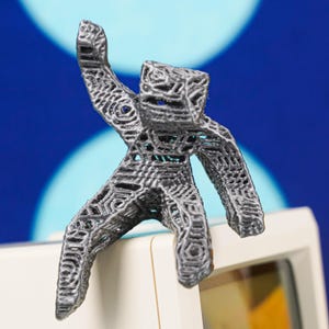 Op de afbeelding: Een donkergrijze, 3D-geprinte figuur van een persoon met opgeheven armen. De figuur heeft een geometrisch, open-lattice ontwerp. Het staat op een wit oppervlak tegen een blauwe achtergrond met witte cirkels.