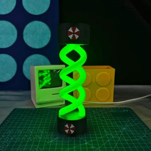 May include: A glowing green DNA-shaped lamp with a black base and top, featuring a red and white umbrella logo. The lamp is set against a backdrop with a green cutting mat, a yellow block, and a retro computer.