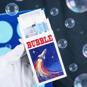 May include: A white rectangular container with the word BUBBLE printed in red letters. The front features a graphic of a space shuttle launching into a dark blue starry sky with a colorful trail. Inside are light blue tubes and a metal cap.