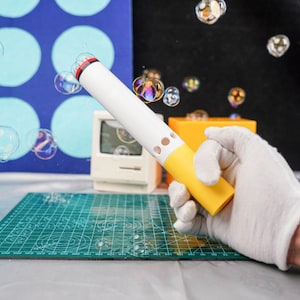 May include: A white and yellow bubble wand with a red accent is held in a gloved hand. The wand is cylindrical with small holes and is creating multiple iridescent bubbles. A miniature computer and other objects are in the background.