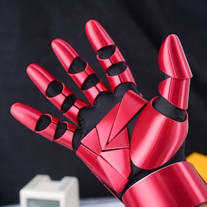May include: A red articulated glove with a black interior. The glove features segmented, metallic-looking plates that cover the fingers and back of the hand. The design suggests a futuristic or robotic aesthetic.