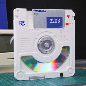 Peut inclure: Un dispositif de stockage de données blanc de style rétro avec un CD visible à travers une fenêtre incurvée. L'appareil porte les inscriptions "TESTODATA HD" et "32GB" sur le devant. Il affiche également "SMD MCD ENTERPRISE CLASS" et "15MB/s".