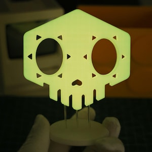 May include: A glowing, light green skull-shaped decoration with large oval eye sockets and triangular accents. The skull is mounted on a white circular base with three metal supports. The design has a geometric, modern aesthetic.