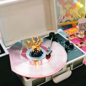 Könnte beinhalten: Ein weißer tragbarer Plattenspieler mit einer rosa Schallplatte. Eine goldfarbene, abstrakte Skulptur sitzt auf der Platte. Der Plattenspieler ist geöffnet und zeigt den Plattenteller und die Bedienelemente. Die Gesamtästhetik ist modern und künstlerisch.