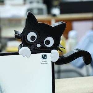 May include: A black cat-shaped desk accessory with white eyes and paws, perched on the corner of a computer monitor. The cat's tail curves around the monitor. The Adobe Photoshop icon is visible on the screen.