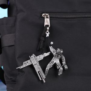 May include: A silver-colored metal charm set featuring a stylized figure and a crossbow design. The charms are attached to a black cord and hang from a backpack zipper.