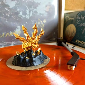 May include: A gold-colored, geometric sculpture resembling a hand with sharp, crystalline fingers, atop a black base. The sculpture sits on an orange vinyl record, with a record player needle in the foreground. The background includes album art.