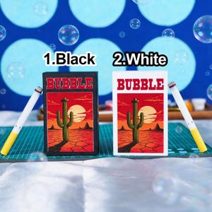 May include: Two rectangular boxes labelled BUBBLE, one black and one white, each featuring a desert illustration with a cactus and a sun. Beside each box is a white and yellow bubble wand. The text 1.Black and 2.White appears above the boxes.