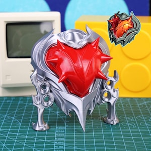 Puede incluir: Una escultura plateada y roja en forma de corazón con una textura puntiaguda. La escultura tiene un aspecto metálico y está colocada sobre una superficie verde. También es visible un pequeño emblema ilustrado del corazón.