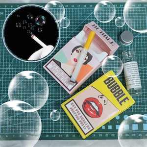 Puede incluir: Un kit de burbujas novedoso con una varita de burbujas con forma de cigarrillo y un diseño de estuche de cigarrillos rosa y amarillo. Las cajas muestran el texto "NOT FOR SMOKING JUST FOR FUN!" y "BUBBLE". También se incluye una pequeña botella de solución de burbujas.