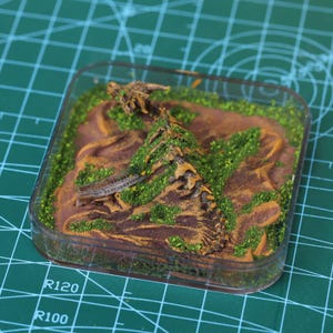 Puede incluir: Un diorama en miniatura en un recipiente de plástico transparente. La escena representa un paisaje marrón texturizado con vegetación verde y una pequeña y detallada figura de dinosaurio. El diorama está sobre un fondo de cuadrícula verde.