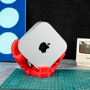 May include: A silver Apple Mac Mini computer with a black Apple logo, held by a bright red crab claw-shaped stand. The stand sits on a green cutting mat. A blue and white painted background is visible.