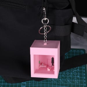 以下が含まれることがあります： 銀色のチェーンと留め金が付いた、ピンク色のミニチュアの部屋のキーホルダー。内部の小さな部屋には家具とフィギュアがあります。キーホルダーは黒いバックパックにぶら下がっています。