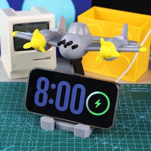 May include: A gray airplane-shaped phone stand with yellow propellers holds a smartphone displaying "8:00" and a charging symbol. The stand is on a green cutting mat, with a yellow box and a retro computer in the background.