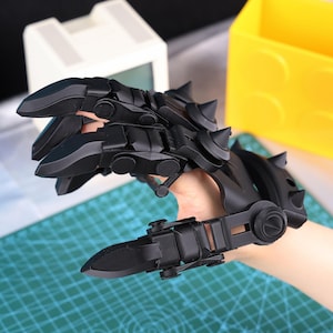 May include: Black articulated claw gauntlet with sharp, curved blades and spiked accents. The gauntlet appears to be made of a hard, durable material. The design is reminiscent of fantasy or science fiction themes.