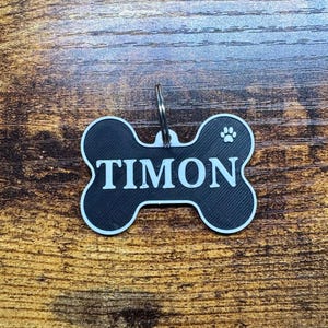 May include: Black and white dog bone-shaped pet tag with the name "TIMON" in bold white letters. A paw print is on the right side. The tag has a silver metal ring for attachment.