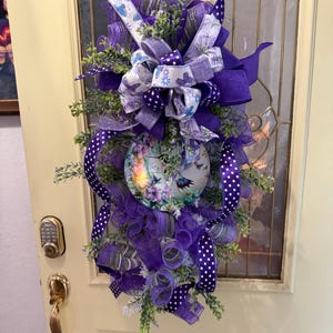 Può includere: Una ghirlanda decorativa a forma di goccia con rete viola, nastri a pois bianchi e motivi floreali. Un cartello circolare centrale mostra un colibrì e dei fiori. Accenti di fogliame verde sono sparsi in tutta la composizione.
