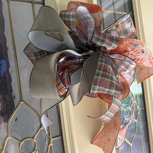 Może przedstawiać: Ozdobna kokarda wykonana z wielu warstw wstążki z drucikiem. Wstążki mają mieszankę jednolitego beżu, szaro-białej kraty oraz pomarańczowych wzorów z jesiennymi nadrukami, w tym dyniami, słonecznikami i jesiennymi liśćmi.