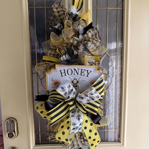 Pode incluir: Uma grinalda decorativa com tema de abelha, com fitas e malha amarelas, pretas e brancas. A grinalda inclui um letreiro que diz "HONEY" com uma ilustração de abelha. Pendurada numa porta creme com painéis de vidro.