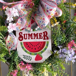 Może przedstawiać: Ozdobny letni wianek z drewnianym znakiem z motywem arbuza i truskawki. Znak głosi "SUMMER" w czerwonych brokatowych literach. Wianek jest ozdobiony kolorowymi kwiatami i kwiatową kokardą.