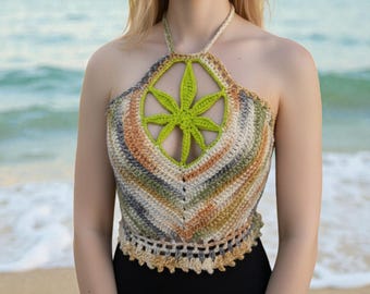 Puede incluir: Top halter de ganchillo hecho a mano con un diseño de hoja de cannabis verde en el centro. El top tiene un patrón de rayas multicolores en tonos beige, verde, naranja y gris. El top se usa en la playa.