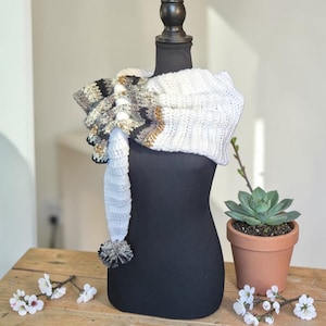May include: A white and patterned crochet scarf with a pom-pom detail, displayed on a black mannequin. The scarf features a mix of white, gray, and gold colors. A small succulent in a terracotta pot and white flowers are in the background.