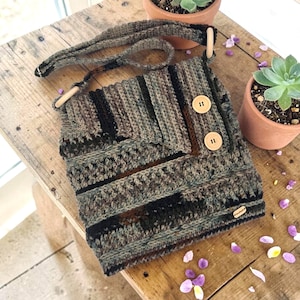 May include: A crocheted crossbody bag featuring a variegated pattern of dark green, brown, and black yarn. The bag has a fold-over flap secured by two light-colored wooden buttons and includes a matching crocheted strap with wooden ring accents.