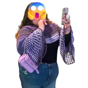 May include: A handmade crochet shawl in shades of purple and gray, featuring bell sleeves and an open weave design. A small, quilted lavender purse hangs from the shawl. The person is wearing blue jeans and a black top.