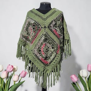May include: A handmade, olive green crochet poncho with a V-neck and fringe. The poncho features a geometric pattern with accents of pink and gray. The garment is displayed on a black mannequin against a white background.