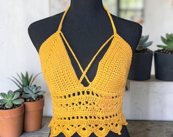 Puede incluir: Top halter de ganchillo amarillo mostaza con un diseño entrecruzado en el busto y un dobladillo festoneado. El top hecho a mano se exhibe en un maniquí negro. Es una prenda de verano.