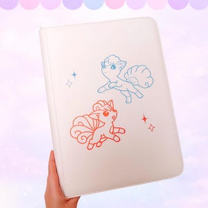 May include: A white, zippered binder featuring blue and orange cartoon illustrations of two fox-like creatures. The binder has a clean, minimalist design with a focus on the playful character art and star accents.