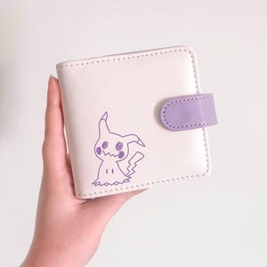 May include: A square, off-white bi-fold wallet featuring a purple line-art illustration of the character Mimikyu on the front cover. The wallet includes a purple snap-button closure tab on the right side and has visible stitching along the edges.