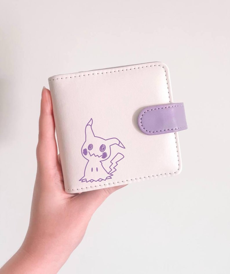 May include: A square, off-white bi-fold wallet featuring a purple line-art illustration of the character Mimikyu on the front cover. The wallet includes a purple snap-button closure tab on the right side and has visible stitching along the edges.