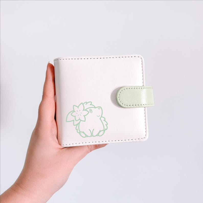 May include: A white bi-fold wallet featuring a light green snap closure and a light green line art illustration of a small creature with a flower on its back. The wallet has a smooth, clean finish and a compact, square-shaped design.