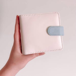 May include: A square, pale pink bi-fold wallet featuring a smooth, matte finish and visible stitching along the edges. The wallet is secured with a small, rounded light blue tab closure that snaps onto the front panel of the wallet.