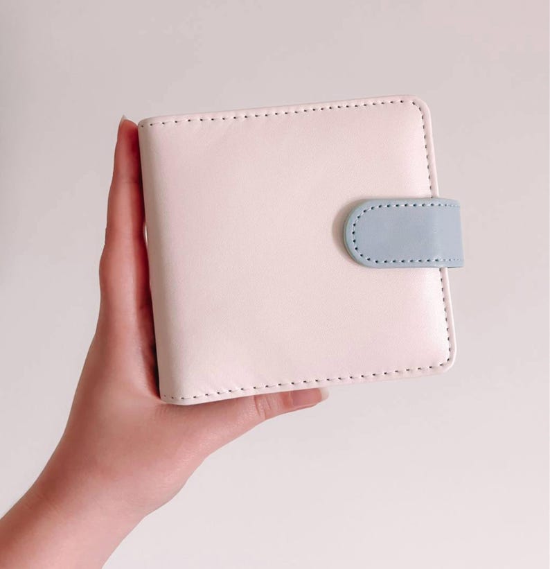 May include: A square, pale pink bi-fold wallet featuring a smooth, matte finish and visible stitching along the edges. The wallet is secured with a small, rounded light blue tab closure that snaps onto the front panel of the wallet.